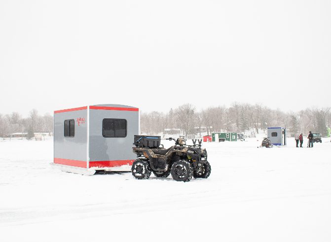 orange slayer skid house on ice with ice shanty town behind it towed by a 4 wheeler