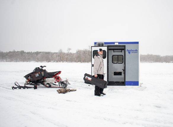 Fisherman loading a skid house with black bag with a snowmobile parked nearby on ice