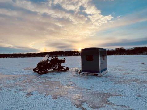 Stryker skid house on ice with snowmobile with sunset behind it