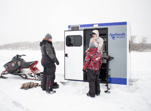 two men and a child talking in front of an ice house on ice with snowmobile nearby